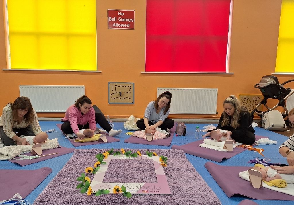 a photo of a 4 women with babies on yoga mats in front of them. In the middle is a soft rug with a selfie frame in the middle which has lots of yellow flowers.