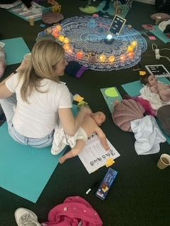 a photo of a yoga mat with a basket of items used in baby massage such as oil, wipes and a colourful scart. There is also a round plaque which states "my first baby massage"